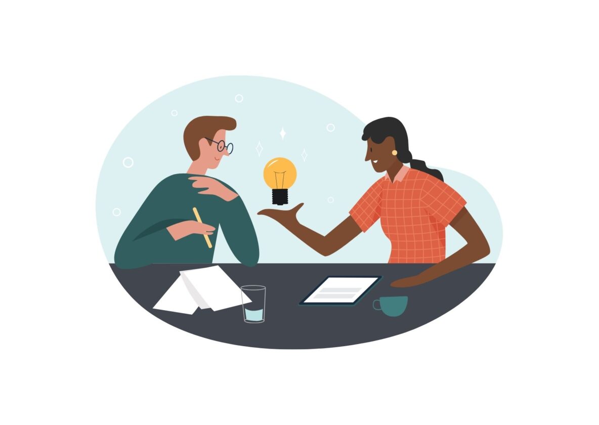 two individuals sitting together at a table with one holding out a lightbulb as if to share an idea