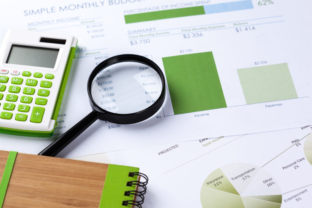 A close-up of financial documents with green charts and graphs, a magnifying glass, a green calculator, and a spiral notebook placed on top.