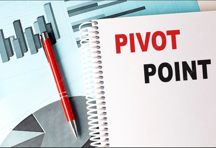 A spiral notebook with the words "PIVOT POINT" written in bold red and black letters, placed on top of charts and graphs, with a red pen resting nearby