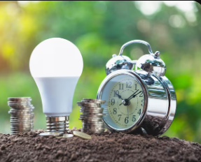 Stacks of coins, an LED light bulb, and a silver alarm clock placed on soil with a blurred green outdoor background, symbolizing energy savings and time management