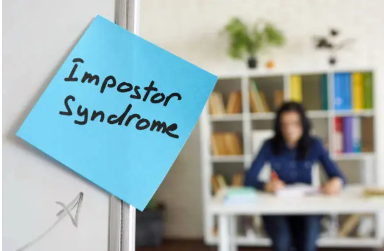 Sticky note reading “Impostor Syndrome” on a glass surface, with a blurred person working at a desk in the background.