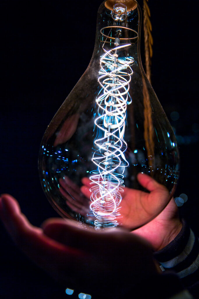A pair of hands gently holding a large clear light bulb with a bright, glowing spiral sparkling filament inside, set against a dark background.