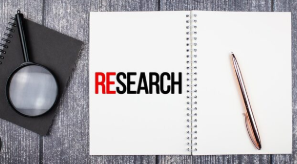 Open notebook with the word “RESEARCH,” a pen, and a magnifying glass on a wooden desk.