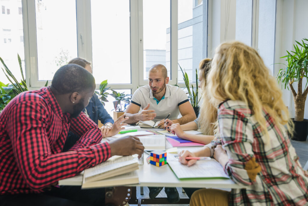 A group of adults sitting around a table in a bright office, focused on a collaborative activity with colorful blocks and papers, suggesting a team brainstorming or problem-solving session.