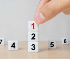 A hand places a numbered block labeled “1” on top of blocks labeled “2” and “3,” with additional numbered blocks scattered around a table